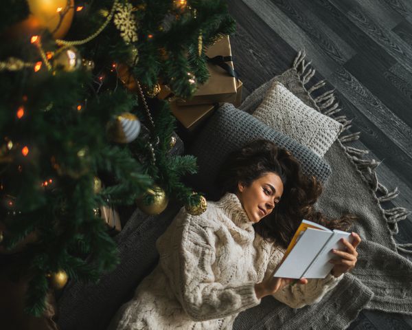 一名女子躺在圣诞树下的枕头和毯子上看书